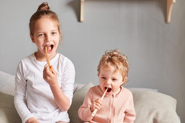 Two little children sitting on the bed in their bedroom with toothbrushes in hand getting ready to brush their teeth after waking up in the morning