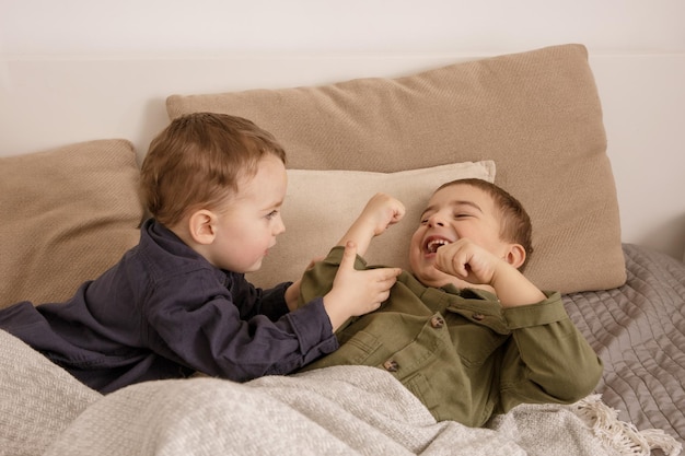 Two little and cute caucasian boys playing together on the bed at home. Interior and clothes in natural earth colors. Cozy environment. Children having fun, two brothers kittle each other.