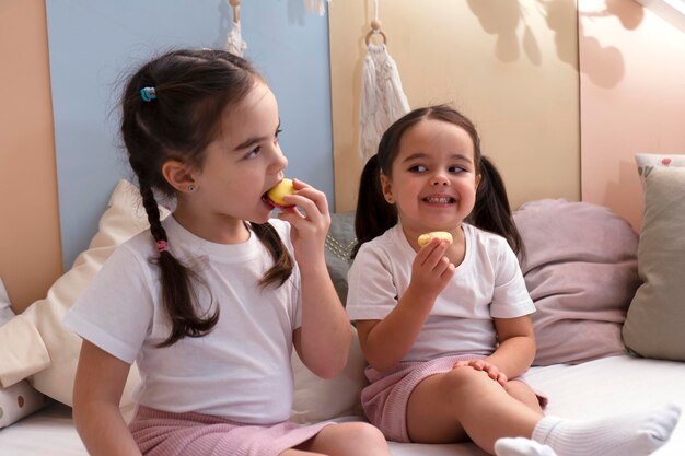 Photo two little cute siblings girls sitting on the bed eating cake macaroon happy moment concept