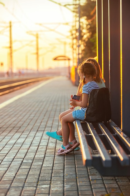 Two little girl tourist sisters sit on the bench of the railway station and look at the phone