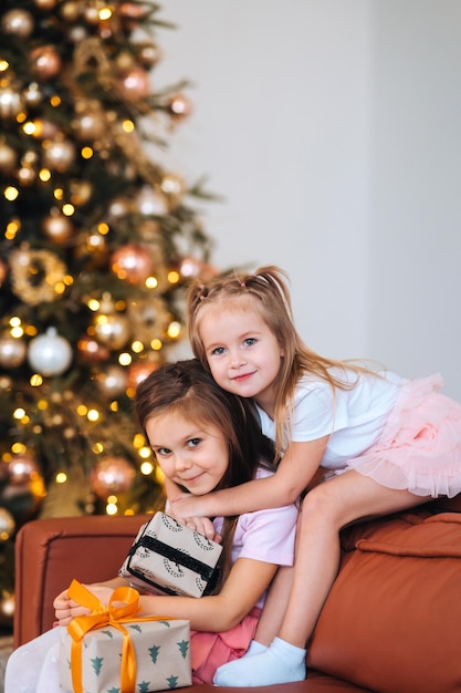 Two little girls are playing with new years gifts by the tree at sofa