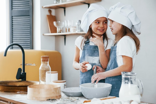Две маленькие девочки в синей форме шеф-повара рассказывают секреты друг другу, готовя еду на кухне