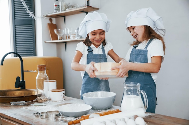 Two little girls in blue chef uniform working with flour by using sieve on the kitchen