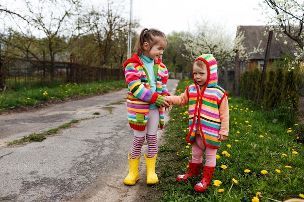 Two little girls in bright clothes hold each other's hands among the yellow dandelions. Happy childhood.