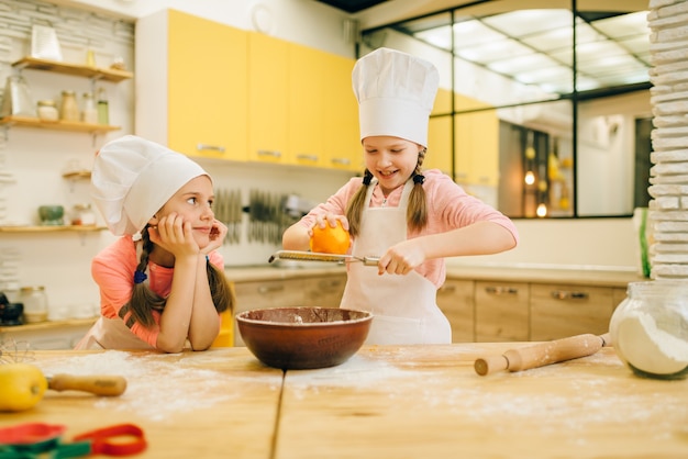 Two little girls cooks in caps rubs orange to the bowl