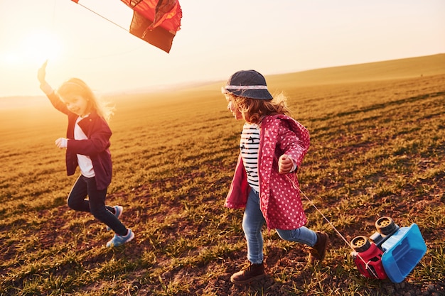 Two little girls friends have fun together with kite and toy car on the field at sunny daytime