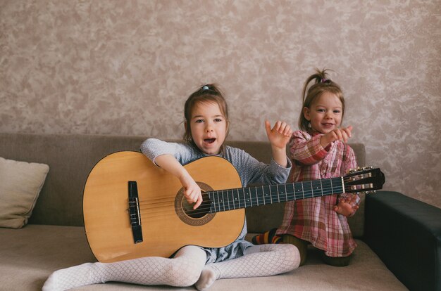 Two little girls learn to play the guitar sing songs sitting at home on the couch