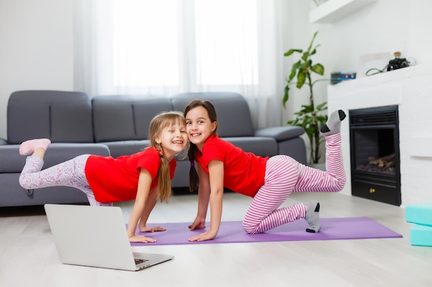 two little girls practicing yoga, stretching, fitness by video on notebook. Distant online education training, aerobic at home. Healthy lifestyle, coronavirus, stay home. Kids sport home quarantine.