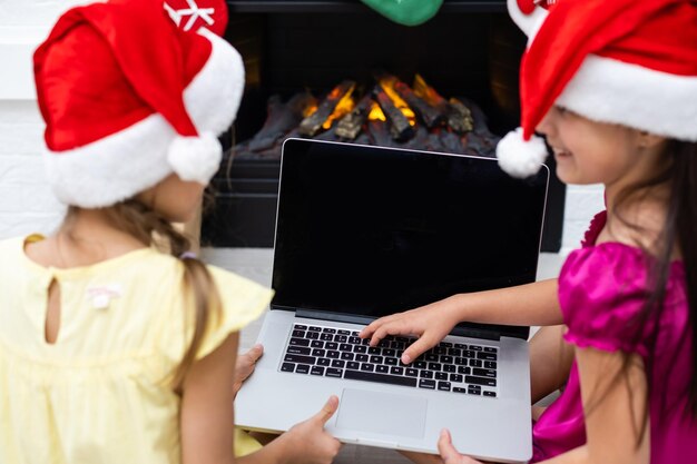 Due bambine, sorelle, amiche, giocano, guardano cartoni animati su un laptop la sera di natale vicino al caminetto