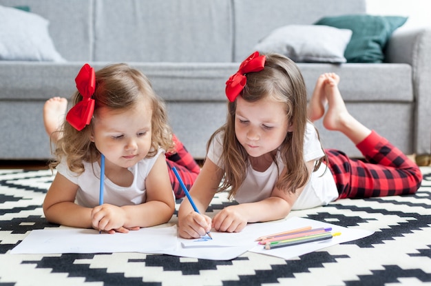 Photo two little girls sisters lie on the floor of the house and draw with colored pencils on paper