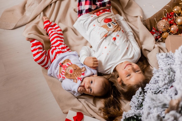 two little girls sisters in New Year's pajamas lie under the Christmas tree at home top view