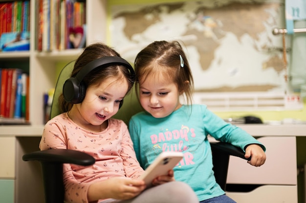 Two little girls sisters wear headphones watching cartoons or kid video on mobile phone