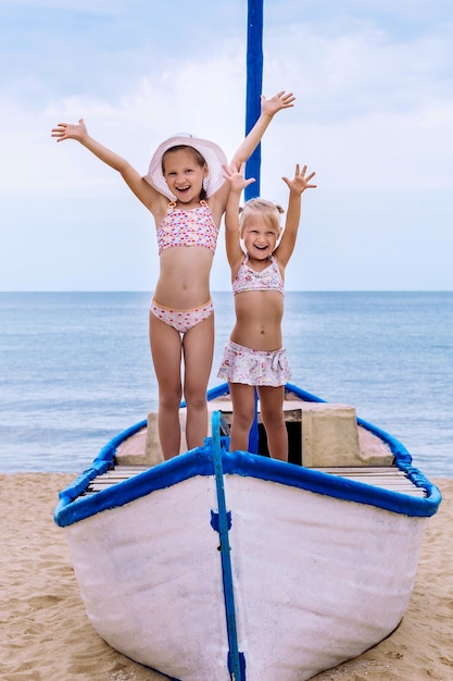 Two little girls in swimwear standing in white and blue boat on sand on sea background raising their hands up