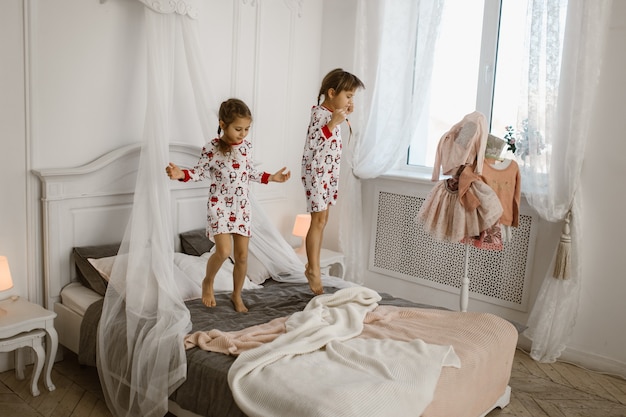 Two little girls in their pajamas are having fun jumping on a bed in a sunlit cozy bedroom . .