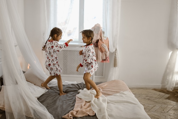 Two little girls in their pajamas are having fun jumping on a bed in a sunlit cozy bedroom . .