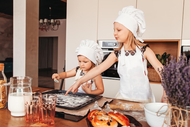 흰색 요리사 모자를 쓴 어린 소녀 두 명이 부엌에서 패스트리를 굽습니다