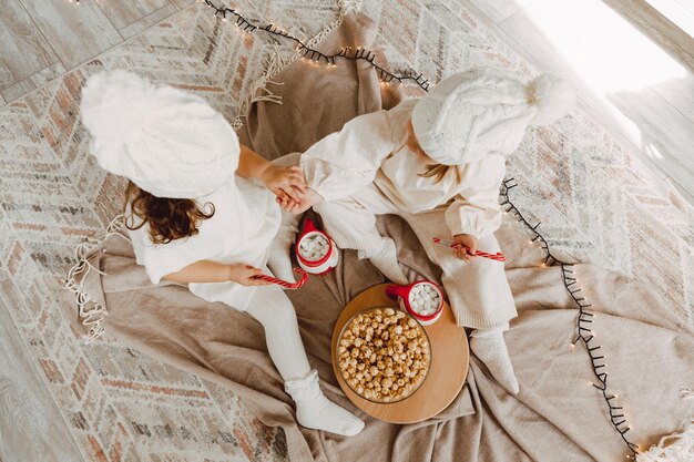 Two little girls in winter knitted hats are sitting on the floor near the Christmas tree and eating popcorn. christmas concept.