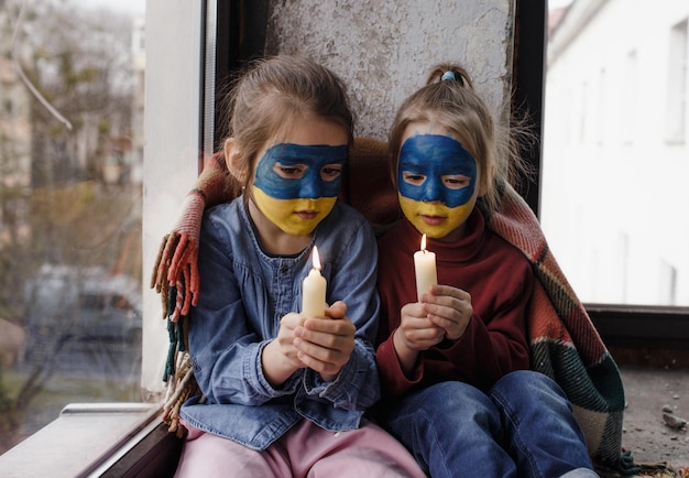 Two little girls with Ukrainian flags painted on their faces are praying for Ukraine holding lighted candles in their hands a gesture of faith and hope Children against war