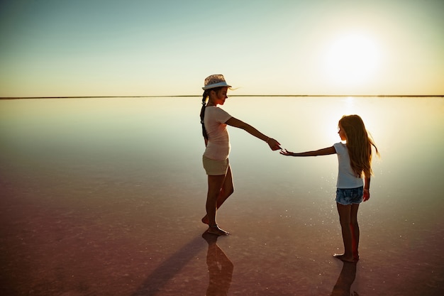 暖かい夕陽を楽しんでいる鏡のピンクの湖に沿って歩いている2人の小さな幸せな姉妹