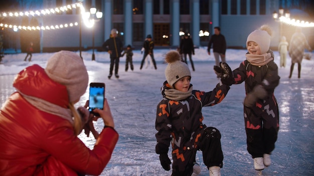 Two little kids on the ice rink  their mother shooting them on the phone camera  kids posing