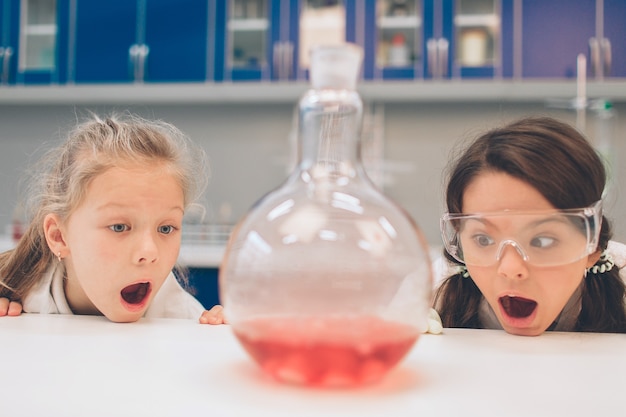 학교 실험실에서 화학을 배우는 실험실 코트에 두 명의 작은 아이. 실험실 또는 화학 캐비닛에서 실험을 만드는 보호 안경의 젊은 과학자. 실험을위한 재료 연구.