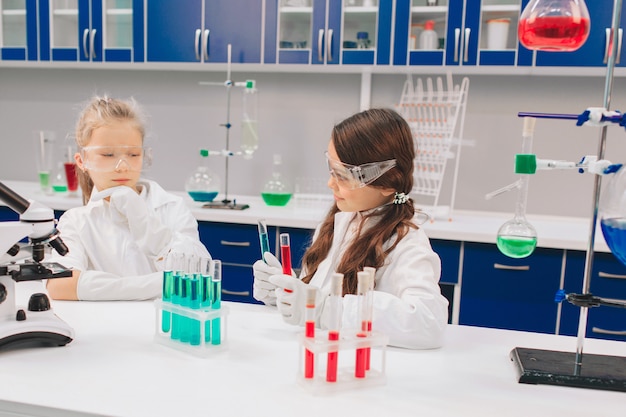 Two little kids in lab coat learning chemistry in school laboratory. Young scientists in protective glasses making experiment in lab or chemical cabinet. Studying ingredients for experiments .