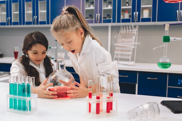 Two little kids in lab coat learning chemistry in school laboratory. Young scientists in protective glasses making experiment in lab or chemical cabinet. Studying ingredients for experiments .