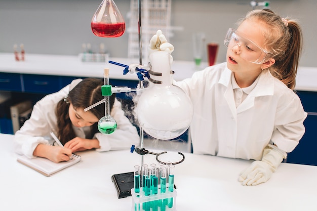 Two little kids in lab coat learning chemistry in school laboratory. Young scientists in protective glasses making experiment in lab or chemical cabinet. Studying ingredients for experiments .