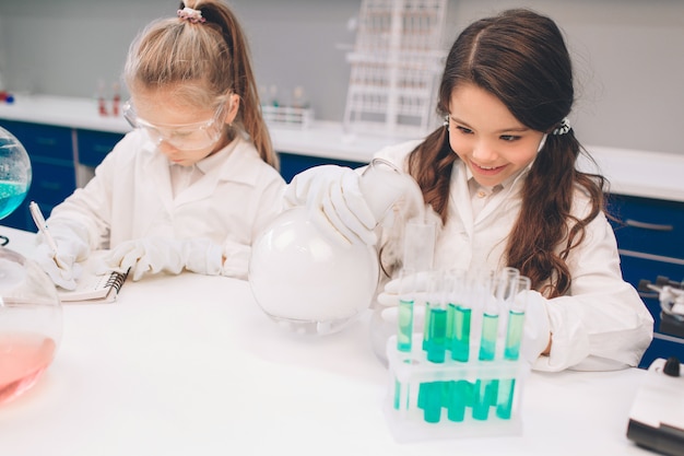 学校の実験室で化学を学ぶ白衣の2人の小さな子供。実験室や化学キャビネットで実験を行う保護メガネの若い科学者。実験の材料を勉強します。