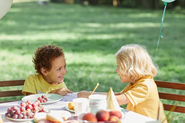 Two little kids at picnic table outdoors decorated with balloons for birthday party in summer copy s...