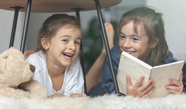 Two little sisters are reading a book lying on the floor on a cozy blanket.