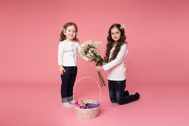 Two little sisters in casual clothes with flowers in their hair standing near a basket of spring flowers