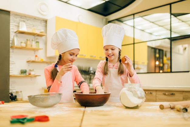 Two little sisters cooks in caps, cookies preparation on the kitchen. Kids cooking pastry, children chefs makes dough, child preparing cake