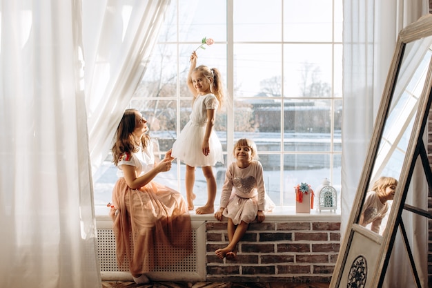 Two little sisters dressed in the beautiful dresses and there young mother sit on the windowsill next to the mirror the winter is outside .