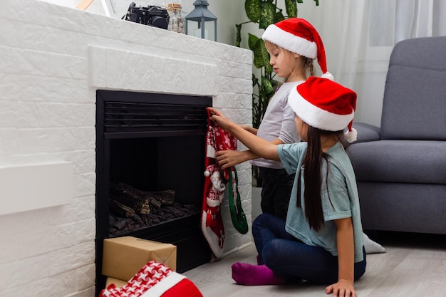 Two little sisters in pajamas having fun New Year's tree with gifts next to the fireplace