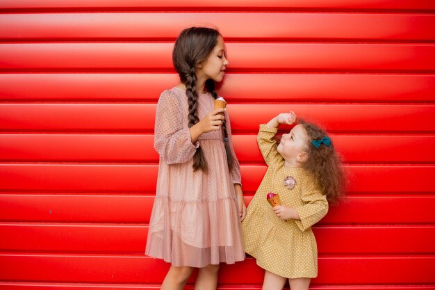 Two little sisters stand at the red fence and eat ice cream. One brunette girl with pigtails, and the second curly baby