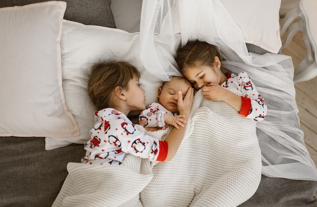 Two little sisters and a tiny brother lie on the bed under the white blanket in the light cozy bedroom . .