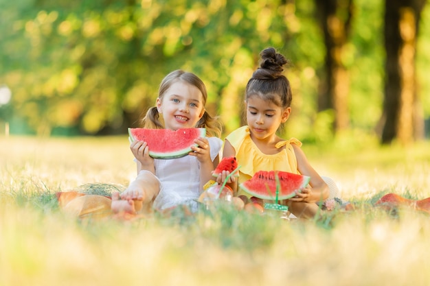 Two little smiling girls playing sitting for picnic and eating piece of watermelon fruit in garden