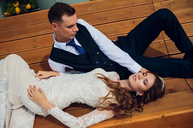 Two lovers sit on a bench, newlyweds crouched to rest in each other's arms during a wedding photo shoot, the bride in a white dress and the groom in a beautiful suit retired in the park.