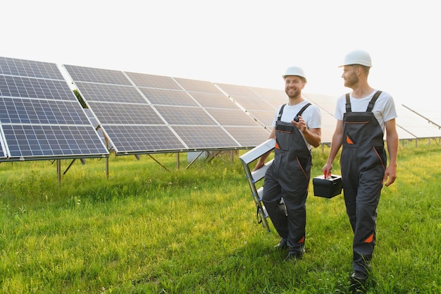 Two male electrician workers walking in between long rows of photovoltaic solar panels and talking about installation of new solar panels