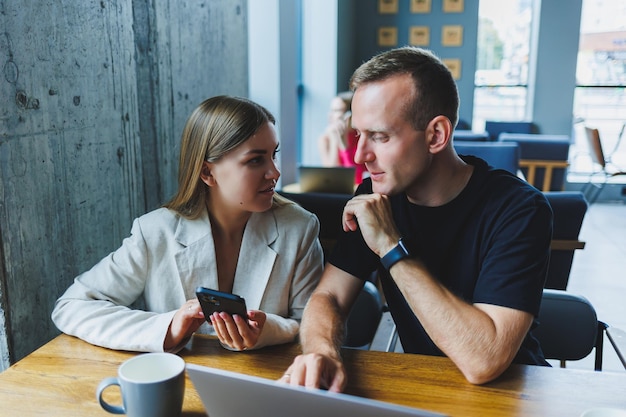 Two male and female colleagues discussing business on laptop
sitting at table in coworking space and talking young male and
female students studying together