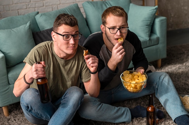 Двое друзей-мужчин пьют пиво с закусками и смотрят спорт по телевизору