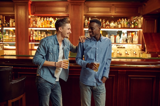 Two male friends poses with glasses of beer at the counter in bar. People relax in pub, night lifestyle, friendship, event celebration