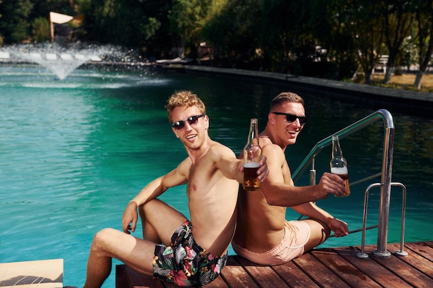 Two male friends sits near water outdoors with drinks in hands