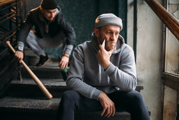 Two male hooligans are sitting on the stairs. Street robbers with baseball bat and knife waiting for victim. Crime concept