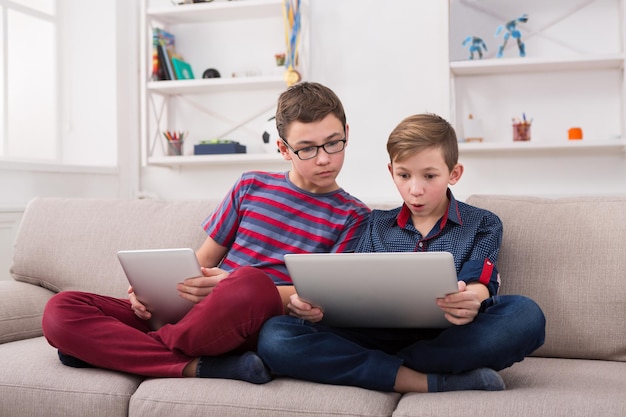 Two male kids with gadgets. Showing each other funny content on laptop and digital tablet on sofa at home. Boy friendship and communication concept