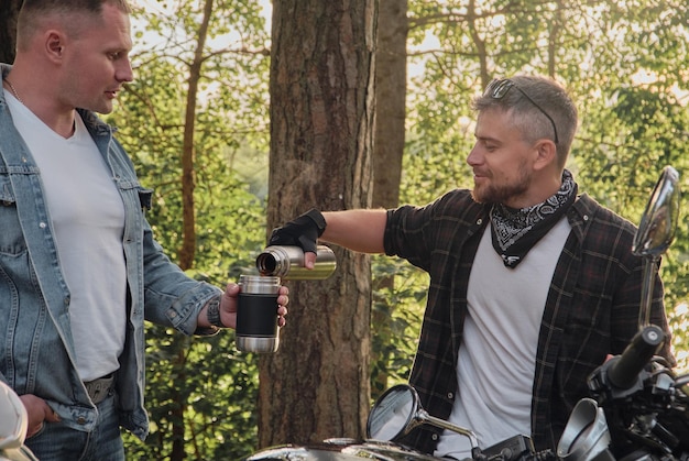 Two man friends of motorcyclists bikers on motorcycles drinking\
tea from a thermos on the side of th