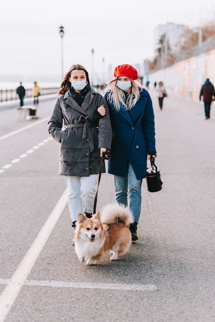 Two masked young women walk a corgi dog together on the street