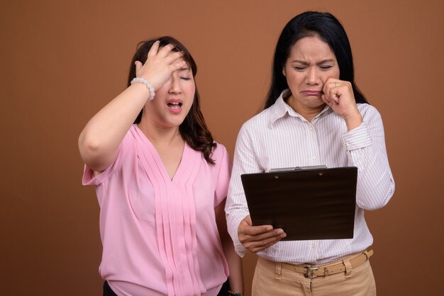 Two mature Asian businesswomen together against brown wall