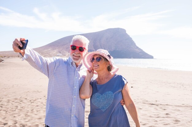 Due persone mature che si fanno un selfie sulla spiaggia sorridenti e si divertono e si divertono - anziani o pensionati che si godono le vacanze o le vacanze all'aperto - giornata di sole in spiaggia